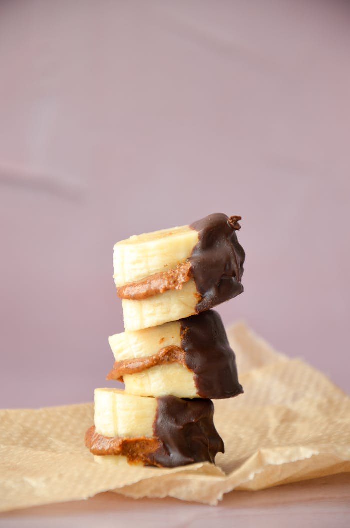 A stack of chocolate-dipped banana slices with nut butter, presented on a rustic paper background.